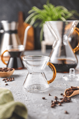 short clip of the 6-cup gold glass handle coffeemaker poring coffee into the gold handle mug, then cream from the cream and sugar set being poured into the mug of coffee. set on a grey kitchen counter with coffee beans and the chettle and a plant in the background,.