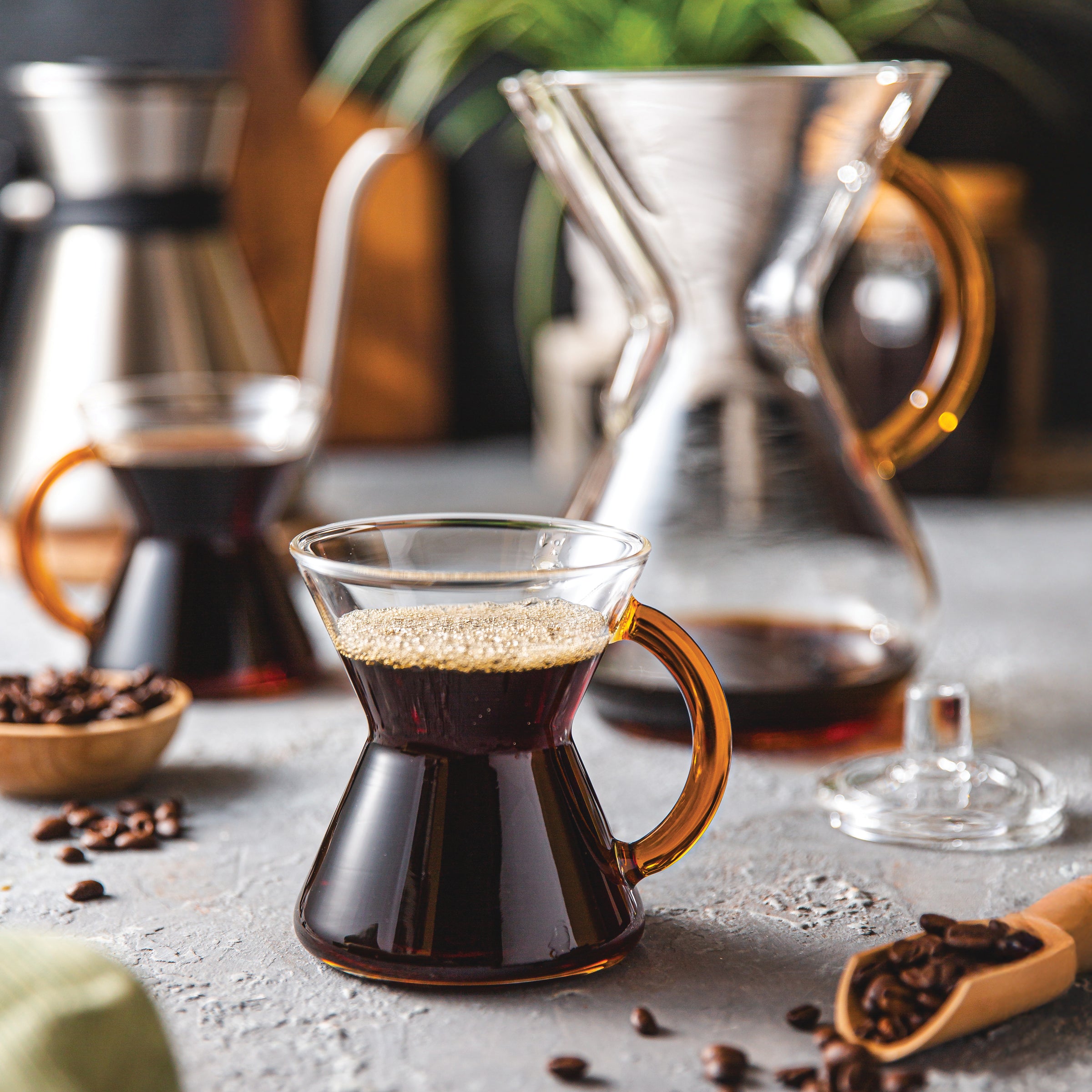 Two gold handle glass mug  and the 6-cup gold handle CHEMEX filled with coffee on a table with coffee beans and the Chettle  in the background.