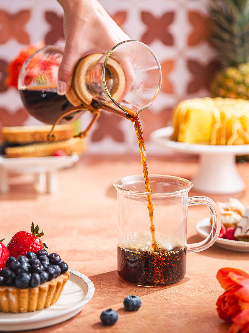 Three Cup Classic CHEMEX pouring coffee into the murray mug, surrounded by colorful fruits and pastries.