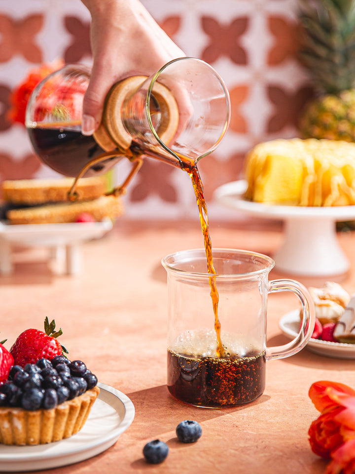 Three Cup Classic CHEMEX pouring coffee into the murray mug, surrounded by colorful fruits and pastries.