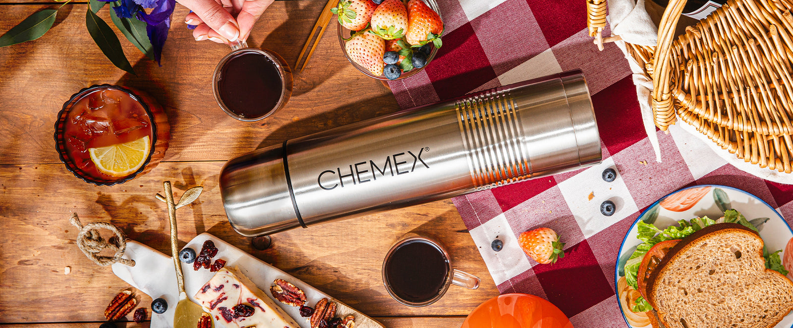 The chemex thermos laying on a picnic table with 2 cups of coffee, surrounded by berries and other picnic foods