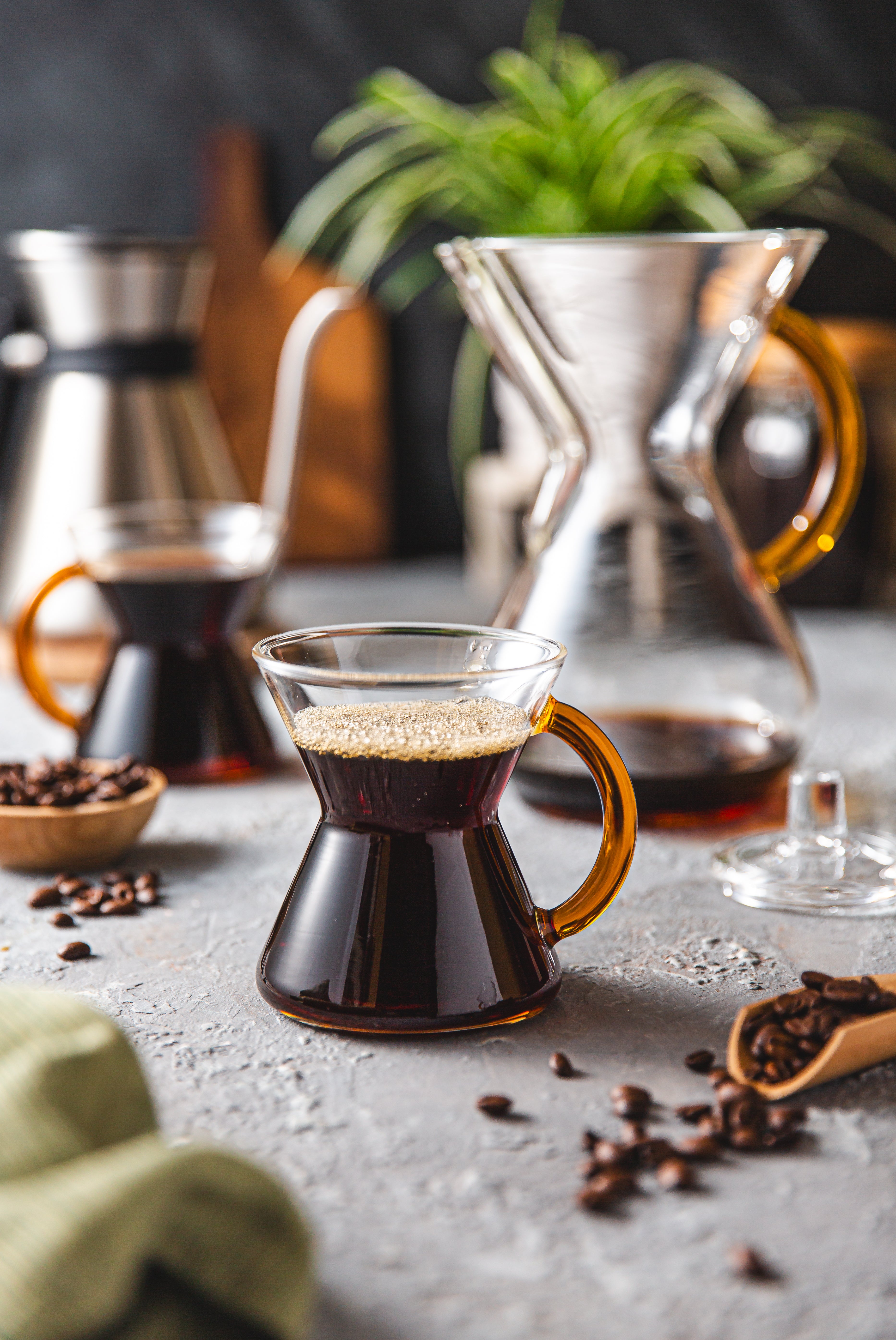 6-cup gold glass handle coffeemaker set which includes 2 mugs filled with coffee on a counter top, coffee beans, a plant and a chettle in the background
