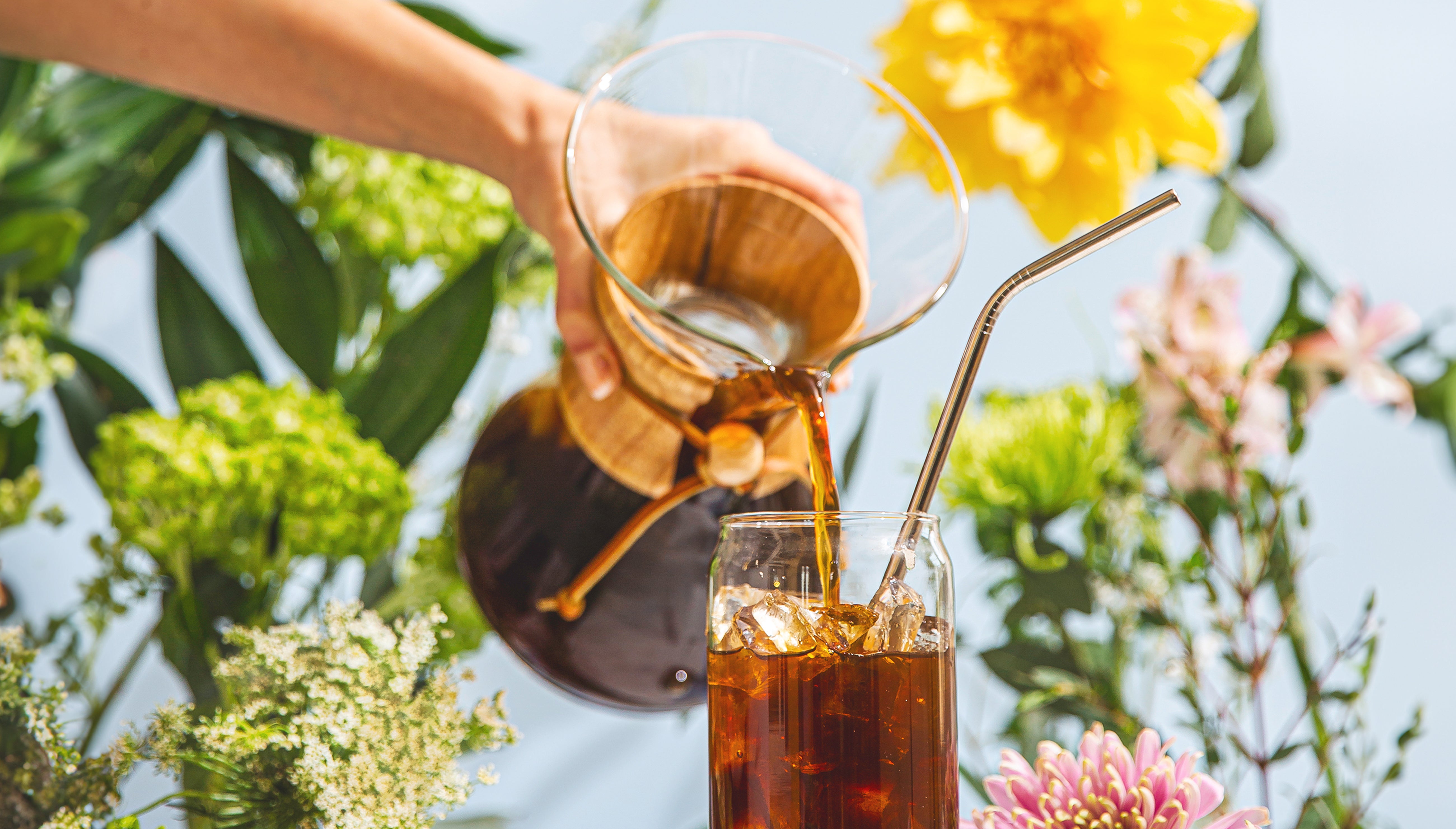 six cup classic coffeemaker pouring iced coffee into a glass with a metal straw, surrounds by flowers and blue sky 