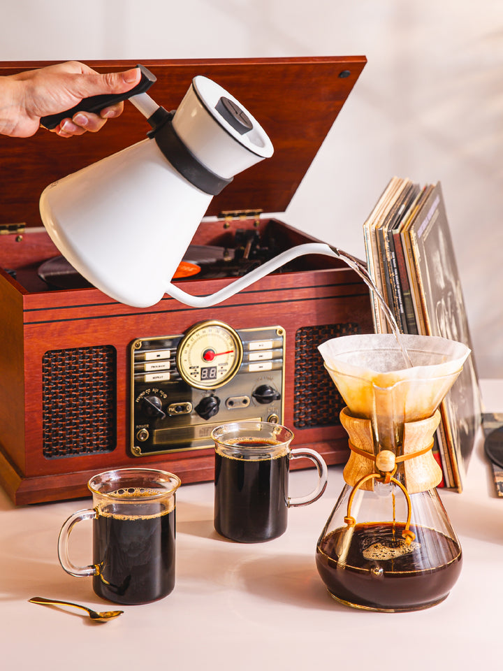 White polished Chettle brewing an eight cup classic CHEMEX pot of coffee. The background is a record played and stack of records.