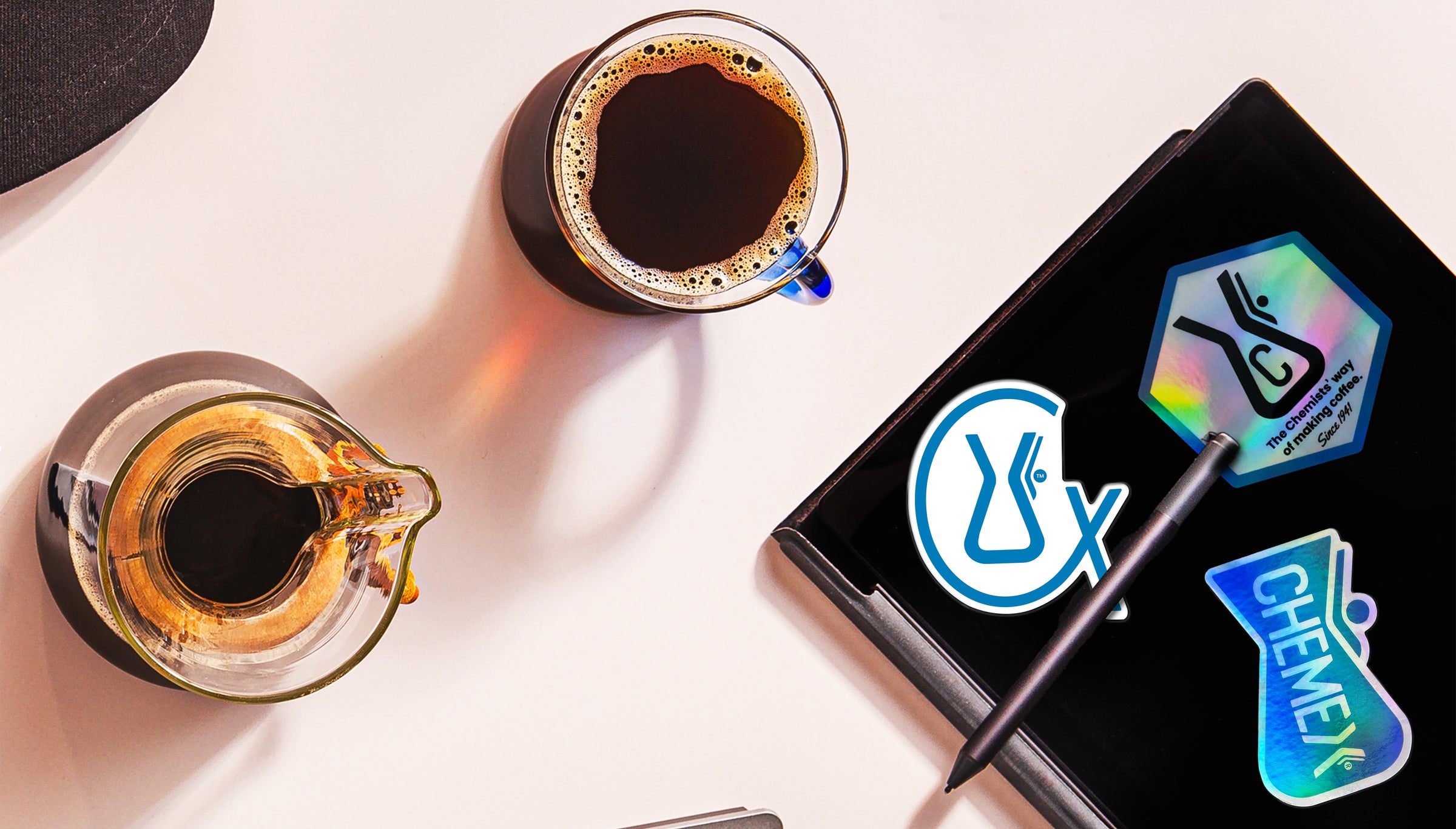 Two cups of coffee with a journal with 3 chemex stickers stuck to its surface.