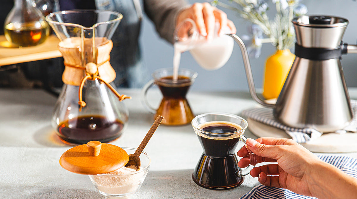 A lifestyle scene of coffee preparation on a light gray countertop, featuring a Chemex pour-over coffee maker, a stainless steel coffee pot, and a hand pouring milk into a glass cup of black coffee.