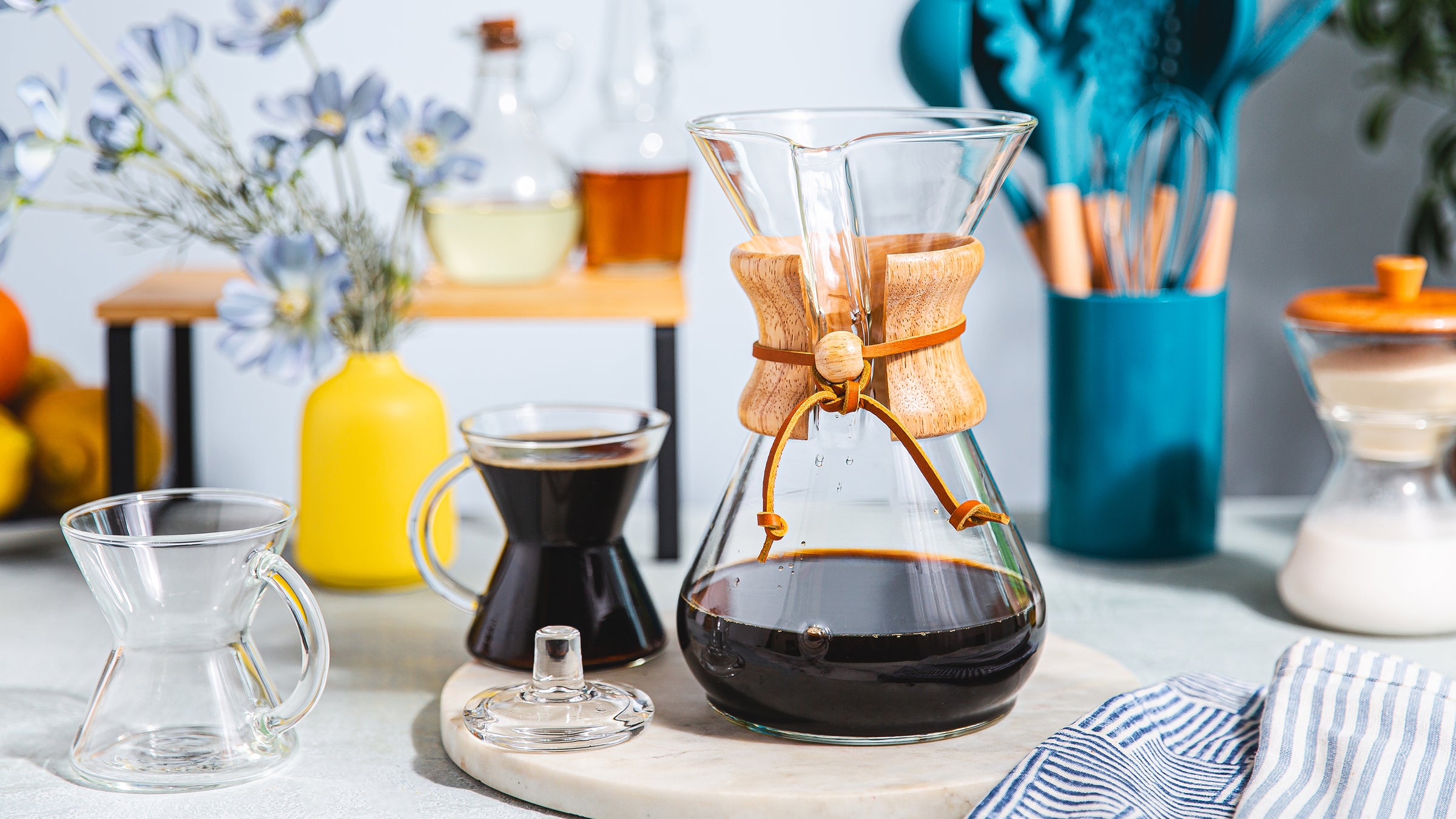 Classic CHEMEX with coffee sitting on a white marble trivet, there is a glass handle mug filled with coffee next to it, another empty glass handle mug and a glass lid. In the background blue cooking utensils and flowers in a yellow vase. 