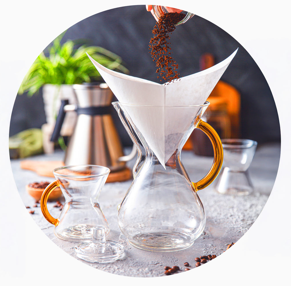 grounds being poured into a filter of a gold handle six cup chemex, a gold handle mug and a glass coffeemaker lid is next to it. the stainless steel chettle is in the background with a green plant.