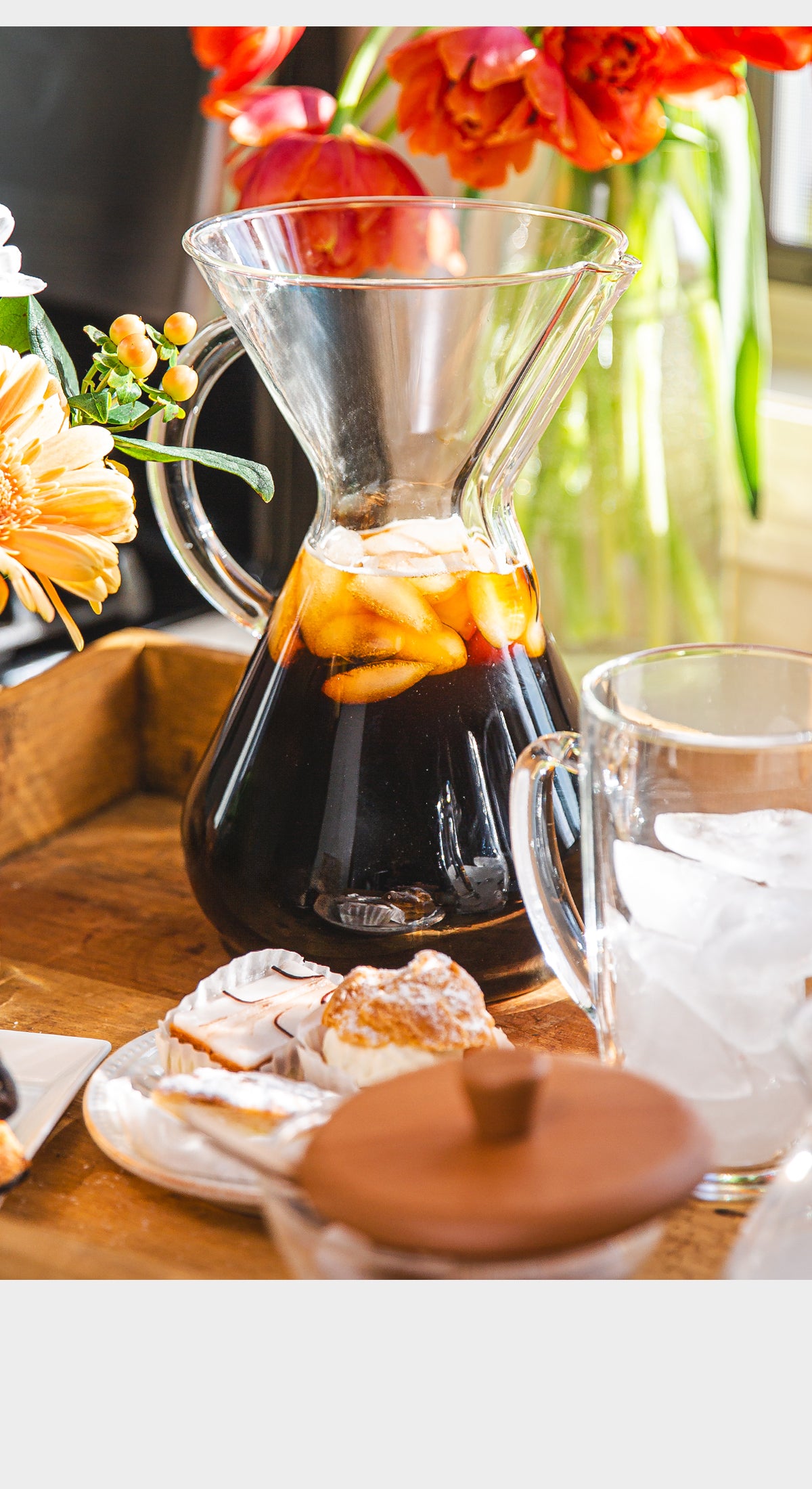 Coffee brewing setup with a glass carafe on a wooden surface, surrounded by flowers and pastries.