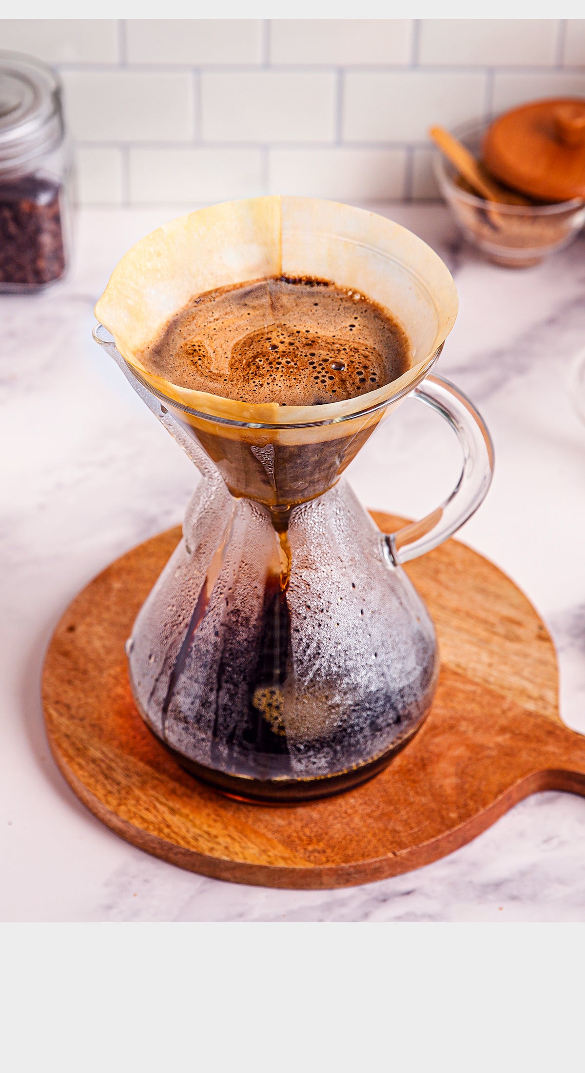 Coffee brewing process with a glass carafe and paper filter on a wooden coaster.