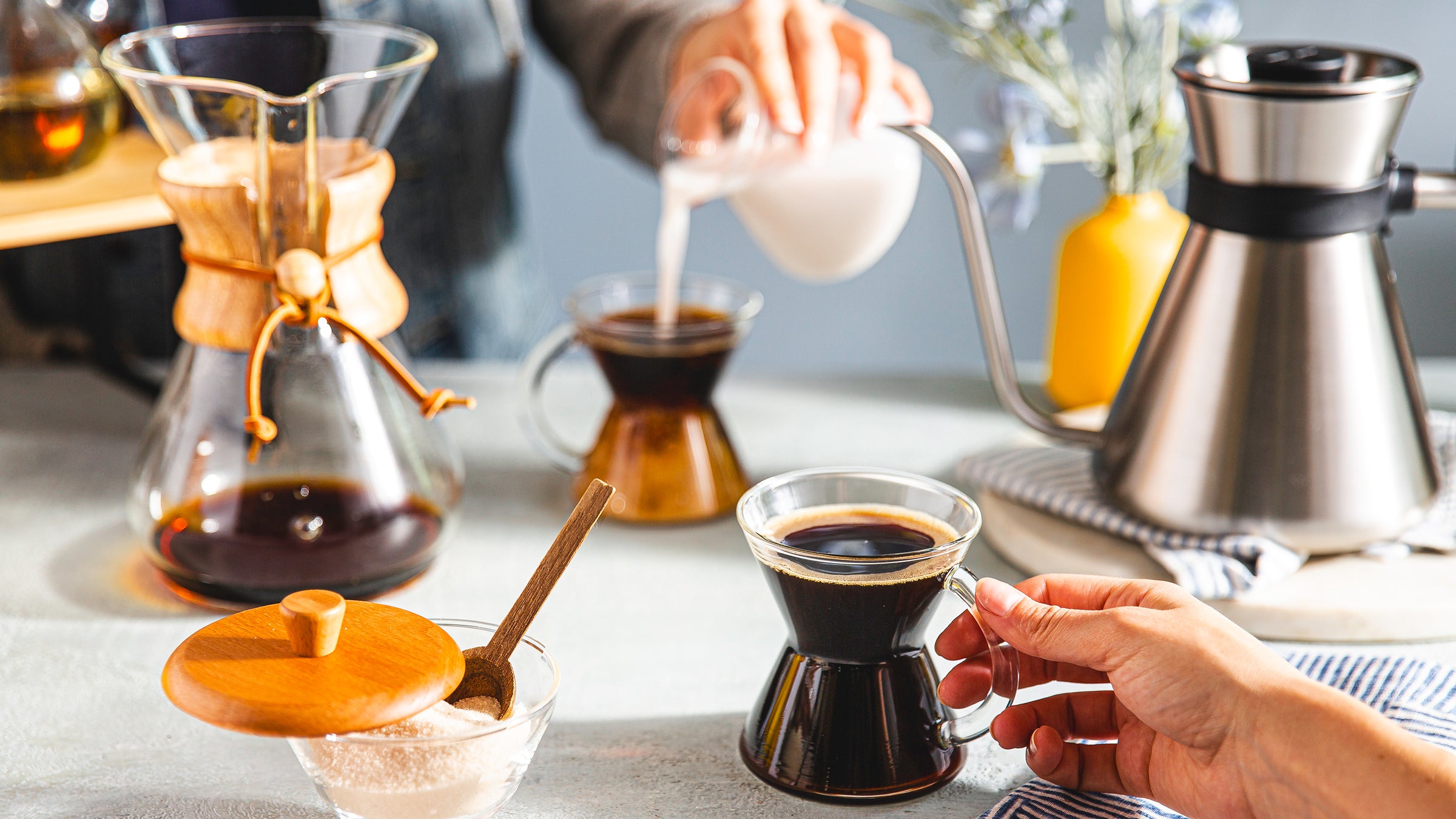 Two people sharing coffee together at a dining room table. There is a chemex coffeemaker, 2 gass handle mugs a cream and sugar set and the chemex chettle.