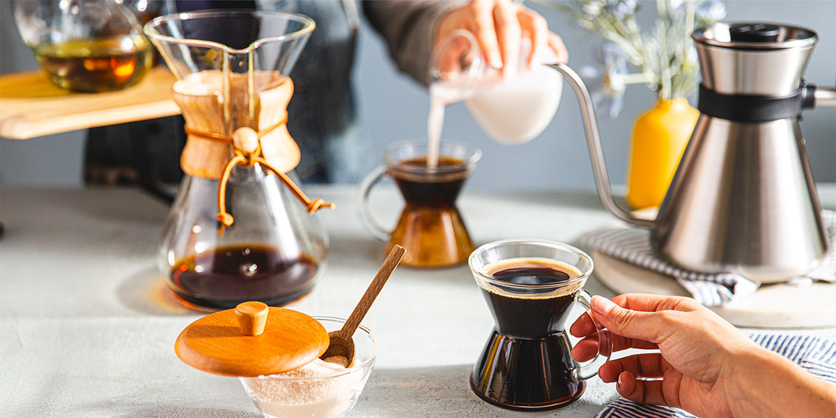 Two people sharing coffee together at a dining room table. There is a chemex coffeemaker, 2 gass handle mugs a cream and sugar set and the chemex chettle.
