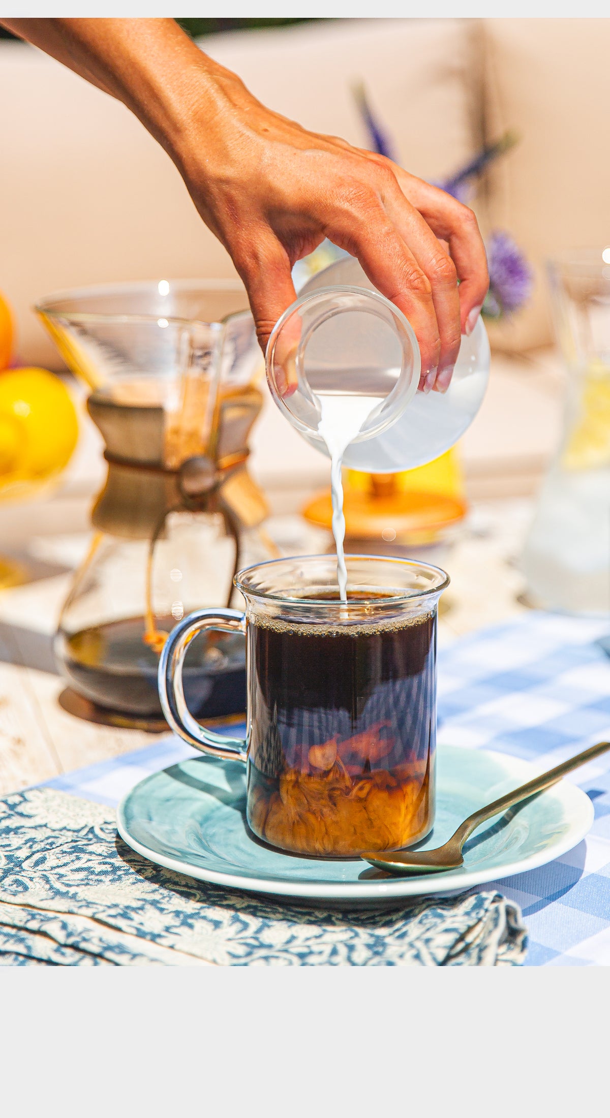 Person pouring milk into a cup of coffee on a table with a checkered tablecloth.